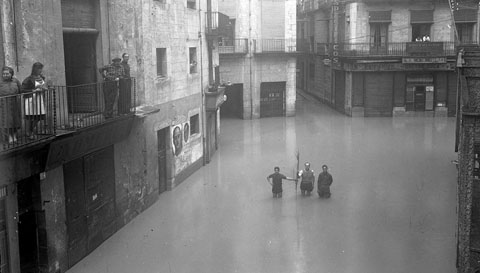 Carrer de la Cort Reial i pla&ccedil;a de l'Oli inundats. A la part central, Daniel Ros Mundet acompanyat de dos nois. Les persones ens donen una idea del nivell on arribaren les aig&uuml;es. A l'esquerra, s'observen cartells del General Franco i de Primo de Rivera. 1940