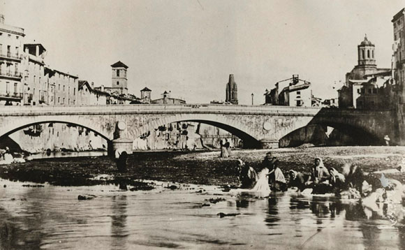 Dones rentant la roba al riu Onyar, davant del pont de Pedra. A l'esquerra, el campanar de les Bernardes i al seu costat, el del convent de Santa Clara. Al centre, entre els arcs del pont de Pedra, s'entreveu l'antiga palanca de les Peixateries Velles que seria substitu&iuml;da pel pont constru&iuml;t per Eiffel. 1870-1873