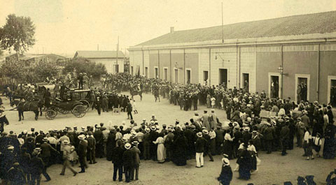 Visita del rei Alfons XIII a la ciutat de Girona. Gent aplegada a la pla&ccedil;a del carril, davant l'estaci&oacute; per rebre el monarca arribat en tren. S'observen diferents carruatges envoltant el vehicle en que viatja el rei i fot&ograve;grafs enfilats a sobre d'un carruatge prenent fotografies. 1904