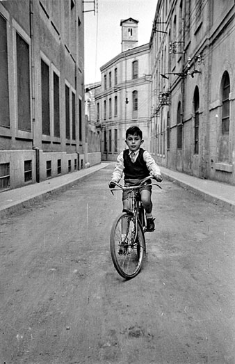 Retrat d'un nen amb bicicleta al carrer de la S&egrave;quia. A banda i banda, la f&agrave;brica Grober. Al fons, la torre de les Aig&uuml;es. 1955