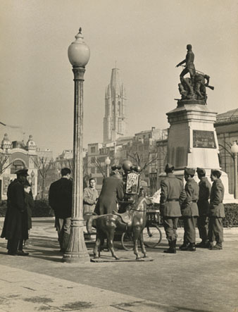 Vista la pla&ccedil;a de la Independ&egrave;ncia. En primer terme davant del monument s'observa un fot&ograve;graf ambulant amb un cavall de joguina envoltat per diverses persones, entre ells quatres soldats. 1957