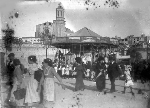 Atraccions de Fires a la pla&ccedil;a de la Independ&egrave;ncia. Al fons, un anunci d'un reconstituent pintat a la paret que tancava la pla&ccedil;a per la banda del riu, la part inferior de la qual encara conserva trets de la fortificaci&oacute; que donava a l'Onyar. El mur desparagu&eacute; el 1914, amb la urbanitzaci&oacute; del passeig Canalejas. 1900
