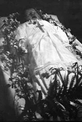 Retrat d'un nad&oacute; mort envoltat de flors i amb una creu. 1908-1929