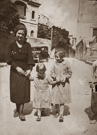 Retrat de d'una dona i dues nenes amb bata escolar al carrer del Portal Nou. A l'esquerra, la casa Gifr&eacute; i a la dreta, l'esgl&eacute;sia de la Merc&egrave;. 1934