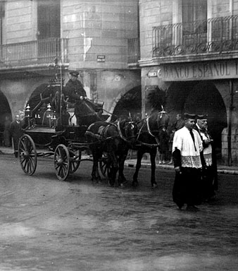 Seguici f&uacute;nebre d'un enterrament al seu pas per la pla&ccedil;a del Vi, amb el carruatge tirat per cavalls engalanats. A la dreta, fa&ccedil;ana del Banc Espanyol de Cr&egrave;dit. 1920