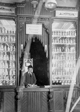 Establiment Ortop&egrave;dia-farm&agrave;cia Doctor Roca. Taulell de la la farm&agrave;cia amb el dependent al darrera i tot de lleixes amb productes farmac&egrave;utics