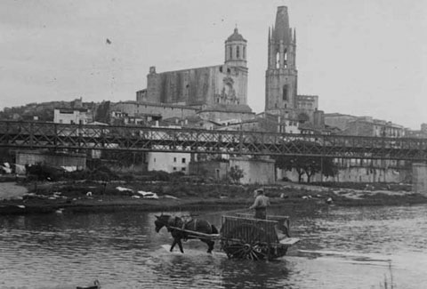 Un carro travessant el riu Ter pel passall&iacute;s del portal de la Barca cobert d'aigua. En primer terme s'observa el pont del ferrocarril. Al fons l'esgl&eacute;sia de Sant Feliu i la Catedral de Girona. 1920-1925