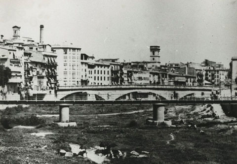 El riu Onyar amb el pont del Pes de la Palla i el pont de Pedra. En primer terme es veuen unes dones rentant al riu. Al fons, el campanar de l'avui desaparegut convent de les Bernardes, la Torre de les Aig&uuml;es i la xemeneia de la Gr&ograve;ber