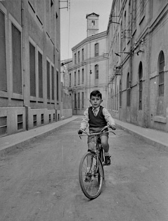 Retrat d'un nen amb bicicleta al carrer S&egrave;quia. A banda i banda, la f&agrave;brica Grober. Al fons, la torre de les Aig&uuml;es. 1955