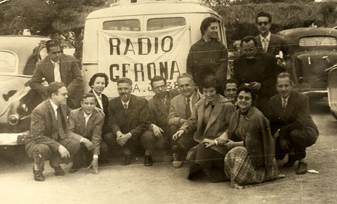 Celebraci&oacute; de la festa de l'Arc&agrave;ngel Gabriel, patr&oacute; dels radiofonistes. Retrat de grup dels treballadors de Radio Gerona EAJ38 davant d'una furgoneta amb una pancarta. S'identifiquen, ajupits, el primer a l'esquerra, Jaume Teixidor, la tercera, Francina Boris i el quart, Emilio Banda. 1955